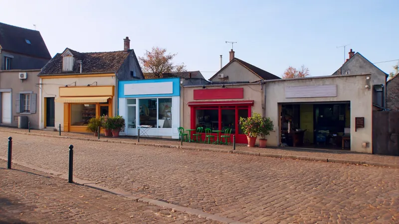 Une rue de petite ville au soleil du matin, avec une boulangerie, un salon de coiffure, un bistrot et un atelier