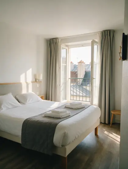 Chambre d'hôtel lumineuse avec un lit fait et une fenêtre ouverte sur les toits